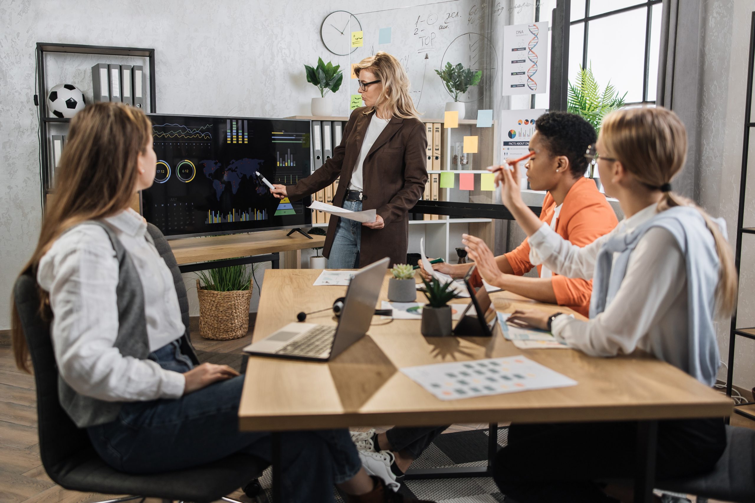 Multiracial female business people having briefing about financial situation at company. Senior woman pointing on monitor with various graphs and charts. Cooperation and brainstorming concept.